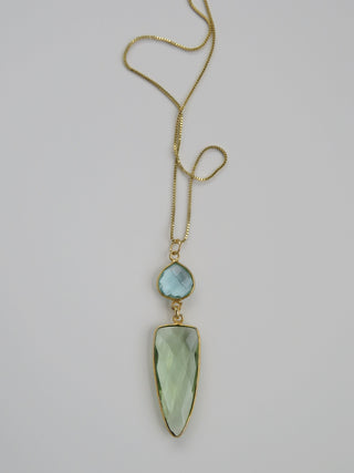 A gold chain necklace featuring a large, faceted light green teardrop gemstone pendant and a smaller round light blue gemstone accent, displayed on a neutral light gray background.